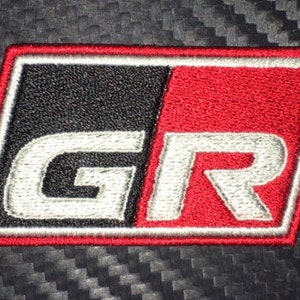 May include: A white embroidered "GR" logo on a black and red patch. The patch has a white border and is rectangular.