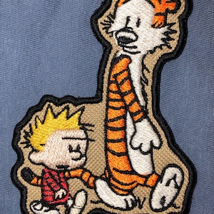 May include: An embroidered patch featuring the cartoon characters Calvin and Hobbes. Calvin is wearing a red shirt and is walking next to Hobbes, a tiger, who is wearing orange and black stripes.