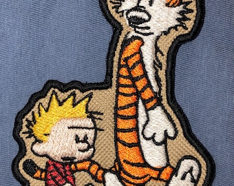 Può includere: Una toppa ricamata con i personaggi dei cartoni animati Calvin e Hobbes. Calvin indossa una camicia rossa e cammina accanto a Hobbes, una tigre, che indossa strisce arancioni e nere.