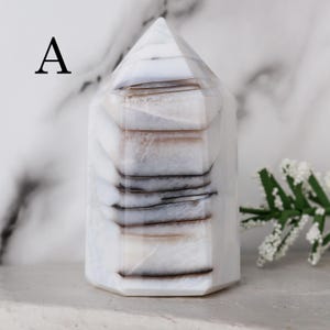 May include: A white and gray agate crystal tower with a hexagonal base and a pointed top. The crystal features bands of gray and brown, creating a layered effect. The letter "A" is in the upper left corner.