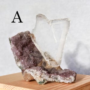 May include: A natural mineral specimen with a large, clear crystal formation on top of a cluster of purple and white crystals. The letter "A" is printed in black in the upper left corner. The mineral sits on a wooden surface.
