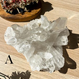 May include: A cluster of clear quartz crystals with multiple points, arranged in a natural formation. The crystals are translucent and reflect light, creating a sparkling effect. The crystals are on a light brown wooden surface.