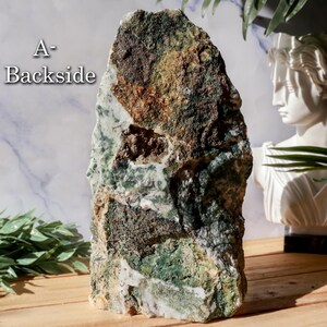 May include: A large, rough-hewn stone with a mix of green, brown, and white hues. The stone has a natural, organic shape and texture. The text "A-Backside" is visible on the upper left side. The stone is displayed on a wooden surface.