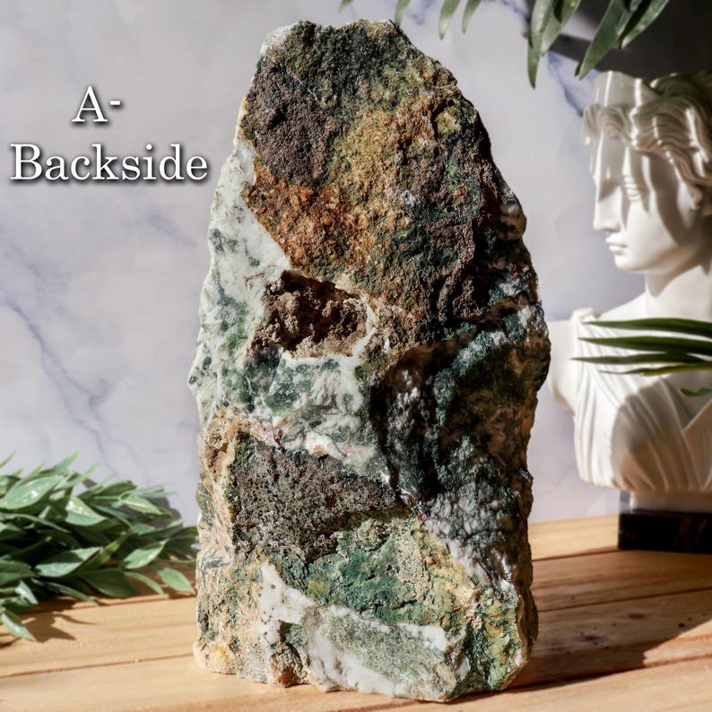 May include: A large, rough-hewn stone with a mix of green, brown, and white hues. The stone has a natural, organic shape and texture. The text "A-Backside" is visible on the upper left side. The stone is displayed on a wooden surface.