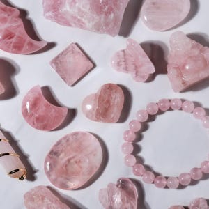 May include: An assortment of pink rose quartz crystals and carved shapes, such as a heart, moon, and square, plus a beaded bracelet. The items are displayed on a white surface, highlighting the natural variations in the stone.