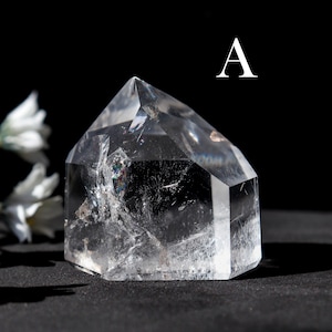 May include: A clear quartz crystal point with a faceted top and sides. The crystal is transparent with internal inclusions and sits on a black surface. The letter "A" is in white in the upper right corner.