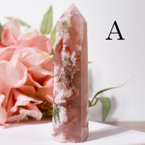 May include: A tall, pink crystal obelisk with white and red veining and green inclusions. The crystal is polished and has a pointed top. The letter "A" is printed in black on the right side of the image. Pink flowers are in the background.