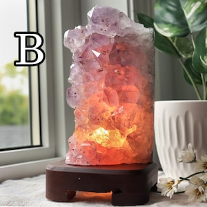 May include: A lit amethyst crystal lamp with a dark wooden base. The crystal is a mix of purple, pink, and orange hues, with a warm glow emanating from within. The letter "B" is in the upper left corner.