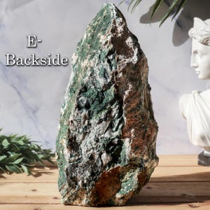 May include: A large, rough-textured stone with a mix of green, white, and brown hues. The stone has an irregular shape and is displayed on a wooden surface. The text "E- Backside" is visible on the stone.