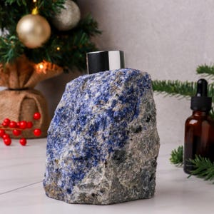 May include: A unique, rock-shaped bottle with a blue and gray stone exterior and a black and silver cap. The bottle is displayed with holiday decorations, including a small Christmas tree and red berries.