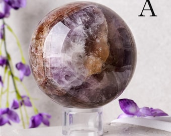 May include: A polished crystal sphere with a mix of purple, brown, and white hues. The sphere is displayed on a clear acrylic stand. The letter "A" is visible in the upper right corner. Decorative purple flowers are in the background.