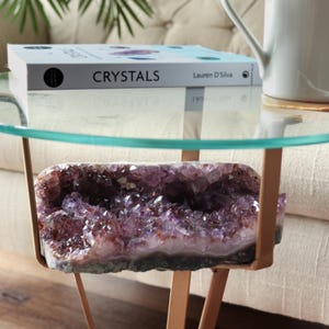 May include: A small, round glass-topped side table with a metal frame. The table features a large amethyst geode as a base. A book titled "Crystals" rests on the glass surface. The geode is a deep purple color.