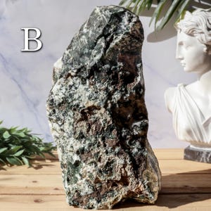May include: A large, rough-hewn stone with a mix of green, brown, and white colors. The stone has a natural, textured surface and is labeled with the letter "B" in white. A white bust and greenery are in the background.