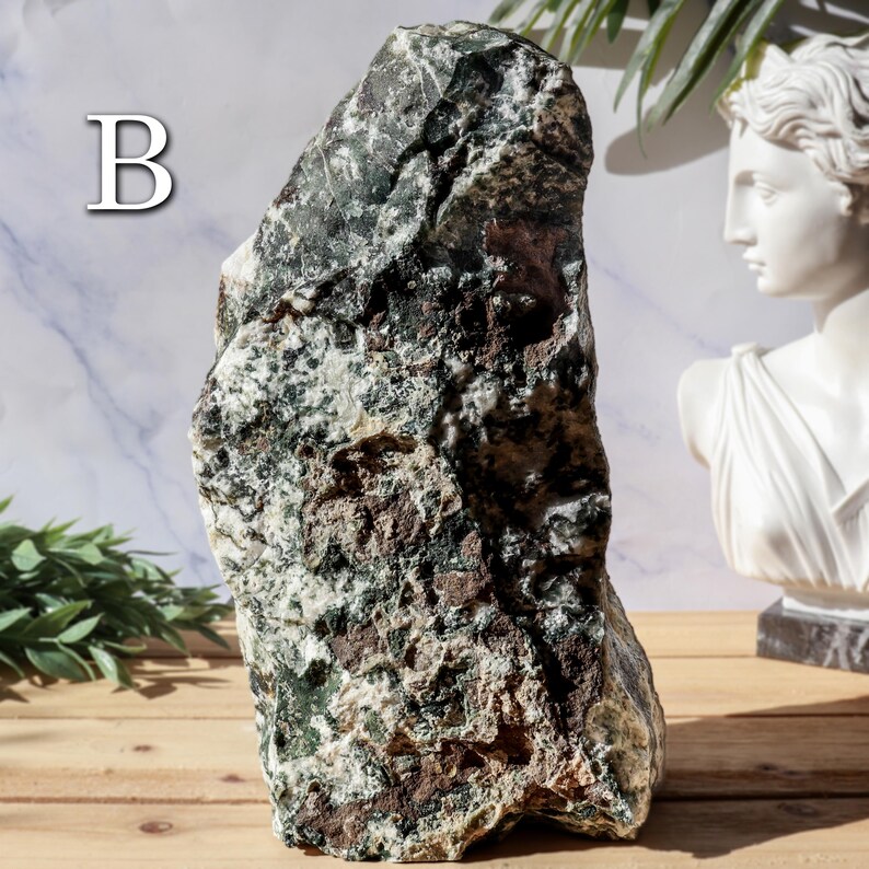 May include: A large, rough-hewn stone with a mix of green, brown, and white colors. The stone has a natural, textured surface and is labeled with the letter "B" in white. A white bust and greenery are in the background.