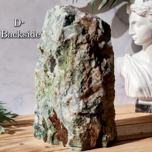 May include: A large, rough-hewn stone with a natural, textured surface. The stone features a mix of colors, including green, brown, white, and red. The text "D- Backside" is visible on the stone. The stone is displayed on a wooden surface.