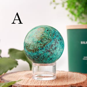 May include: A teal and brown sphere, possibly a decorative stone or crystal, is displayed on a clear acrylic stand. The sphere has a mottled appearance with varying shades of teal and brown. The letter "A" is in the upper left corner. The sphere is on a wooden surface.
