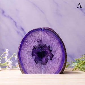May include: A pair of purple agate bookends featuring a natural crystalline geode center with deep violet hues and a polished outer edge. The two halves form a rounded shape, showcasing the raw, jagged texture of the interior quartz crystals.