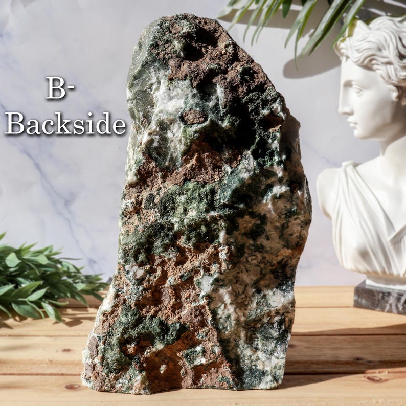 May include: A large, rough-hewn stone with a mix of green, brown, and white hues. The stone has a natural, organic shape and texture. The text "B- Backside" is visible on the stone. A white bust is in the background.