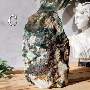 May include: A large, rough-hewn stone with a mix of green, brown, and white colors. The stone has a natural, organic shape and texture. The letter "C" is visible on the left side of the stone. The stone is displayed on a wooden surface.