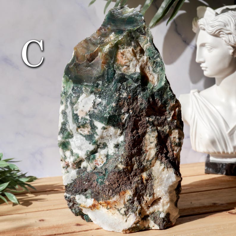 May include: A large, rough-hewn stone with a mix of green, brown, and white colors. The stone has a natural, organic shape and texture. The letter "C" is visible on the left side of the stone. The stone is displayed on a wooden surface.