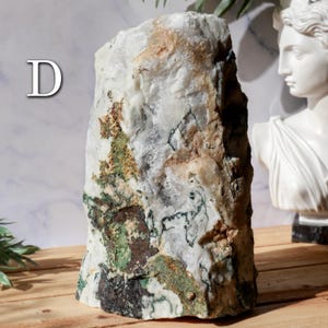 May include: A large, rectangular stone with a rough, textured surface. The stone is primarily white with patches of green, brown, and tan. The letter "D" is visible in the upper left corner. A white bust is in the background.