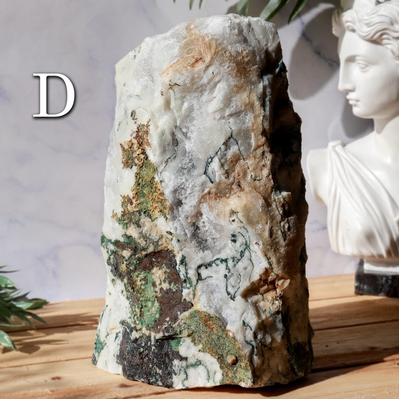 May include: A large, rectangular stone with a rough, textured surface. The stone is primarily white with patches of green, brown, and tan. The letter "D" is visible in the upper left corner. A white bust is in the background.