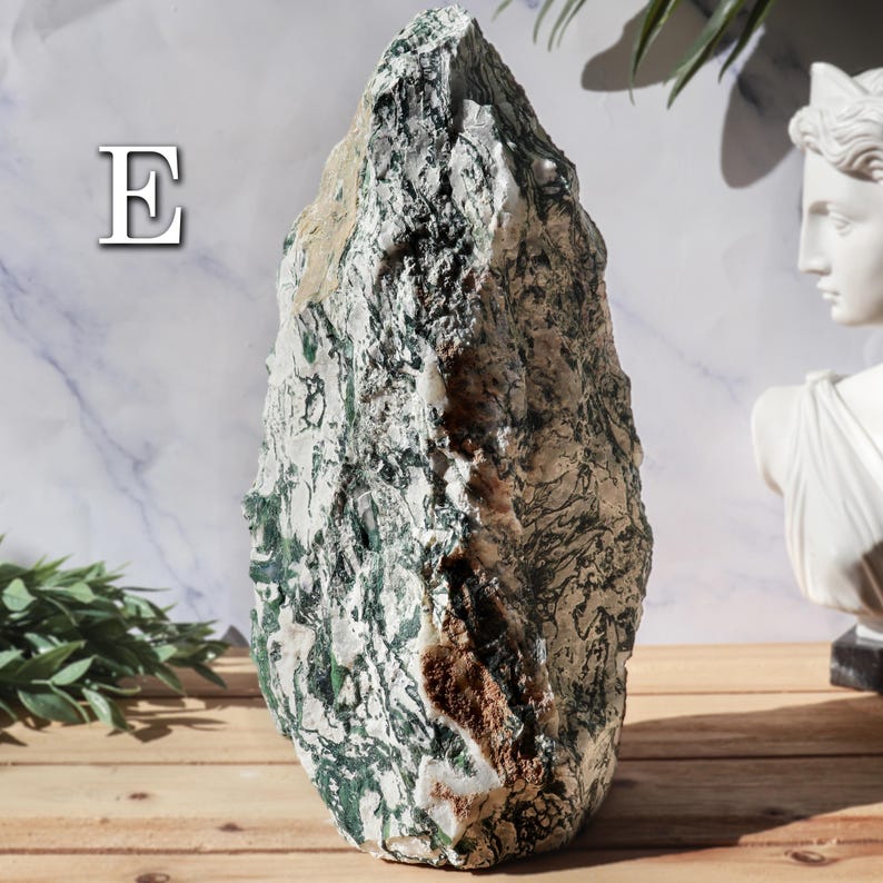 May include: A large, natural moss agate stone with a rough, textured surface. The stone features a mix of white, green, and brown colors, with intricate patterns. The letter "E" is visible in the upper left corner. The stone is displayed on a wooden surface.