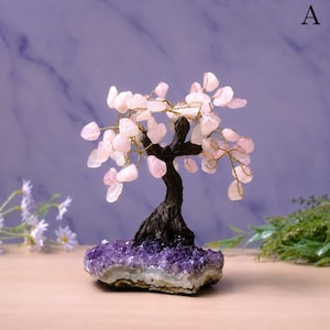 May include: A decorative tree sculpture featuring a dark brown textured trunk and branches made of thin gold wire. The branches are adorned with numerous small, polished light pink rose quartz stones. The base is a cluster of deep purple amethyst crystals.