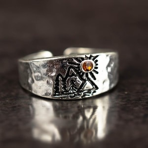 Könnte beinhalten: Ein silberner Ring mit gehämmertem Muster und einem eingravierten Berg- und Sonnendesign. Der Ring hat einen kleinen, runden, orangefarbenen Edelstein. Der Ring ist verstellbar und liegt auf einer dunklen, reflektierenden Oberfläche.