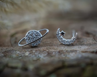 May include: A pair of silver earrings with a planet and a crescent moon design. Both earrings are adorned with small, clear crystals.