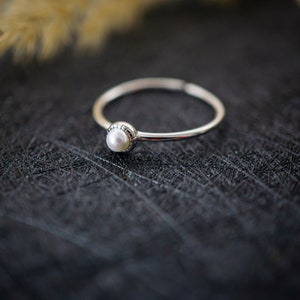 Könnte beinhalten: Ein zarter Silberring mit einem einzelnen weißen Perlmutt, der in einer kleinen, dekorativen Fassung sitzt. Der Ring liegt auf einer schwarzen, strukturierten Oberfläche.
