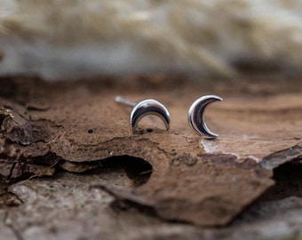 May include: A pair of silver crescent moon stud earrings on a piece of bark.