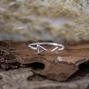 Op de afbeelding: Een zilveren ring met twee driehoeksvormen op de band. De ring ligt op een stuk schors.