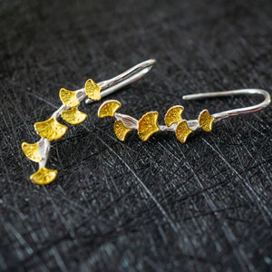 Può includere: Un paio di orecchini d'argento con accenti di foglie di ginkgo dorate. Gli orecchini hanno la forma di un ramo con più foglie.