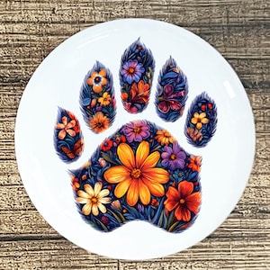 May include: A white ceramic plate featuring a colorful floral paw print design. The paw print is filled with orange, yellow, purple, and red flowers, set against a dark blue background. The plate has a glossy finish and is suitable for decorative use.
