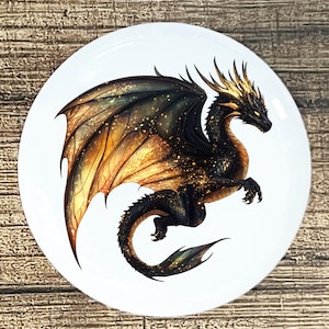 May include: A white ceramic plate featuring a detailed illustration of a black dragon with gold and orange wings. The dragon has a long tail and is set against a white background. The dragon's wings are spread, and the plate has a glossy finish.