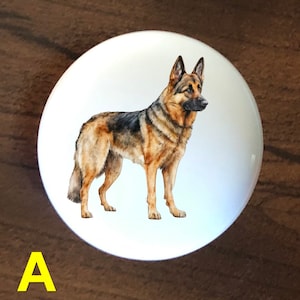 May include: A white ceramic knob with a watercolor illustration of a German Shepherd dog. The dog is standing on all four legs and looking to the right. The background is white.