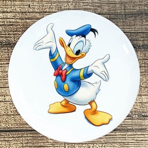 May include: White ceramic plate featuring a colorful illustration of Donald Duck. Donald Duck is depicted in his signature blue sailor suit, bow tie, and hat, with his arms outstretched in a welcoming gesture. The plate has a glossy finish.