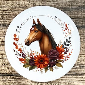May include: White ceramic plate featuring a horse head illustration. The horse is brown with a black mane and a white star on its forehead. The horse is surrounded by a floral wreath with orange, burgundy, and green accents. The plate has a rustic, farmhouse aesthetic.