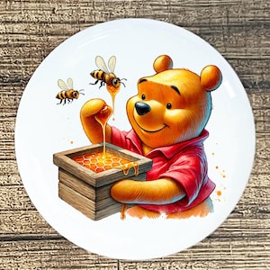 May include: A white ceramic plate featuring a cartoon illustration of a yellow bear in a red shirt, holding a wooden box of honey. Two bees are flying around the bear. The plate has a wooden background.