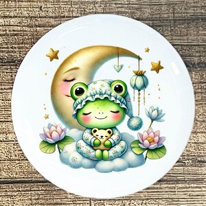 May include: A white ceramic plate featuring a whimsical illustration. A green frog wearing a nightcap and pajamas sits on a cloud, holding a teddy bear. A golden moon and stars are in the background, with lotus flowers and hanging decorations.