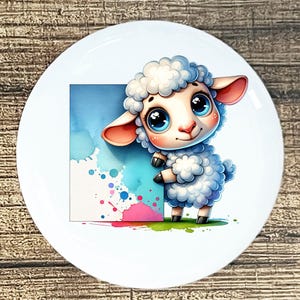 May include: White ceramic plate featuring a cartoon sheep with big blue eyes and fluffy white wool. The sheep is next to a watercolor design with blue, pink, and white splashes. The plate has a glossy finish.