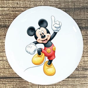 May include: White ceramic plate featuring a colorful illustration of Mickey Mouse. Mickey is depicted in his classic outfit, with red shorts, yellow shoes, and white gloves, pointing upwards with a cheerful expression. The plate has a glossy finish.