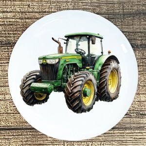 May include: A white ceramic plate featuring a detailed illustration of a green tractor with black tires and yellow accents. The tractor has a cab, a steering wheel, and a smokestack. The plate is round and has a glossy finish.