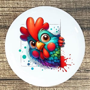 May include: A white ceramic plate featuring a colorful cartoon rooster peeking from behind a white rectangle. The rooster has a red comb, blue and green feathers, and a red wattle. Red paint splatters are on the right side of the plate.