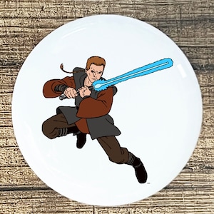 May include: White round ceramic plate featuring a cartoon illustration of a character wielding a blue lightsaber. The character is depicted in a dynamic pose, wearing brown and gray attire. The plate has a glossy finish.