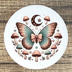 May include: A white ceramic plate featuring a whimsical design. A large butterfly with teal and pink wings is the focal point, adorned with a pink bow. The design includes mushrooms, stars, and a crescent moon, all in soft pastel colors.