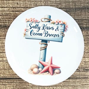 May include: White ceramic plate with a beach-themed design. A light blue wooden sign with the text "Salty Kisses & Ocean Breezes" is surrounded by pink flowers. A pink starfish, seashells, and sand complete the coastal scene.