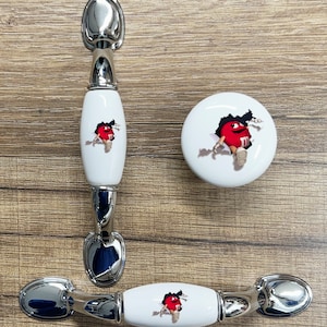 May include: Three white ceramic cabinet knobs with silver metal accents. The knobs feature a red M&M character peeking out of a hole in the ceramic.