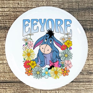 May include: A white round button with a blue and purple cartoon donkey surrounded by colorful flowers. The text "EEYORE" is written in a blue and purple gradient at the top of the button.